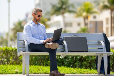 Kendine güvenen işadamı bankta dizüstü bilgisayarı ve parktaki telefonuyla oturuyor. Zengin iş adamı. Olgun iş adamı tatilin tadını çıkarıyor. İş adamı parktaki bankta mola veriyor. Yaz işleri.