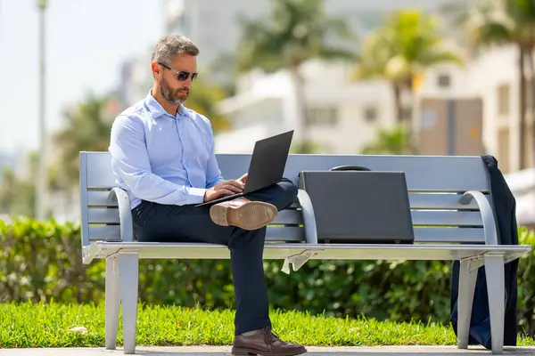 Kendine güvenen işadamı bankta dizüstü bilgisayarı ve parktaki telefonuyla oturuyor. Zengin iş adamı. Olgun iş adamı tatilin tadını çıkarıyor. İş adamı parktaki bankta mola veriyor. Yaz işleri.