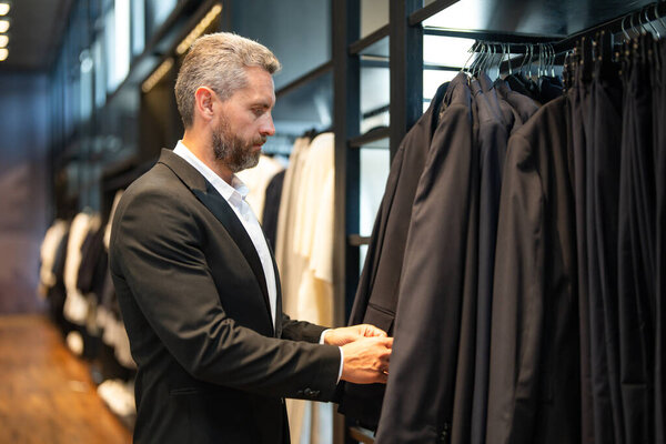 Пожилой мужчина в костюме в бутике костюмов. Бизнесмен уверенно выбирает элегантный костюм в магазине. Модный клиент рассматривает куртку во время шопинга формального костюма. Очаровательная модель в классическом наряде