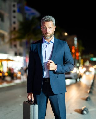 Gece vakti evrak çantalı bir iş adamı yürüyor. Şehirde takım elbiseli zengin bir adam. Lüks arabanın yanında bir iş adamı. Şehir ışıklarının yanında yürüyen bir iş adamı. İş stili. Lüks ortamda zengin bir işadamı