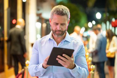 Gece sokakta tablet kullanan bir iş adamı. İş adamı dışarıda dijital tablet üzerinde çalışmayı planlıyor. Neon şehir ışıkları altında bir tablet üzerinde gezinen bir iş adamı. Serbest çalışan bir iş adamı. Gece işi.