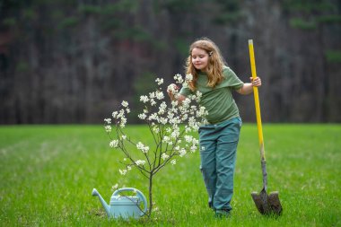 Bahçe bahçesinde çocuk bahçıvanı ağaç dikiyor. Bitkilerin bahar etkinliği. Çocuk kürek ve su bidonuyla çalışıyor. Çocuk meyve ağacı dikmiş. Bahçıvan çocuklar. Çocuk yetiştirme bitkisi ve ağaç bakımı