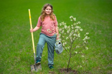 Çocuk çiftçisi, bahar bahçesinde çiçek ve sulama ağacı. Bitkilerin bahar etkinliği. Çocuk kürek ve su bidonuyla çalışıyor. Çocuk meyve ağacı dikmiş. Bahçıvan çocuklar. Çocuk yetiştirme bitkisi ve ağaç bakımı