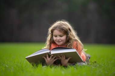 Çimlerin üzerinde kitap okuyan bir çocuk. Çocuk açık havada kitap okumaktan zevk alıyor. Güneşli bir günde çayırda kitapla ders çalışan bir çocuk. Doğada öğrenme. Öğrenci çocuk kitap okuyor. Çocuk açık havada öğreniyor