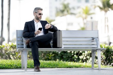 Kendine güvenen bir iş adamı bankta dizüstü bilgisayarı ve parktaki telefonuyla oturuyor. Zengin iş adamı. Olgun iş adamı tatilin tadını çıkarıyor. İş adamı parktaki bankta mola veriyor. Yaz işleri.