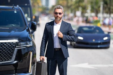 Dışarıdaki iş adamı portresi. Zengin işadamı başarının tadını çıkarıyor. Şık takım elbiseli lüks iş adamı. Pahalı arabanın önündeki zengin adam. Lüks iş seyahati. Klasik takım elbiseli, kendinden emin zengin bir iş adamı.