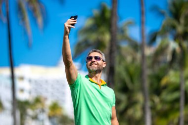 Turist palmiye ağacının yanında selfie çeker. Adam yaz tatilinde selfie çekiyor. Gezgin, tropikal bir yerde selfie çekiyor. Palmiye ağaçlı selfie tropikal titreşimleri yakalar.