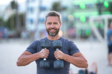 Sportif adam dambılları kaldırıyor. Formda bir adam, dışarıda spor salonunda mankafalarla egzersiz yapıyor. Sporcu dambılları kaldırıyor. Halterli orta yaşlı adam eğitimi. Halterli açık hava fitness antrenmanı.