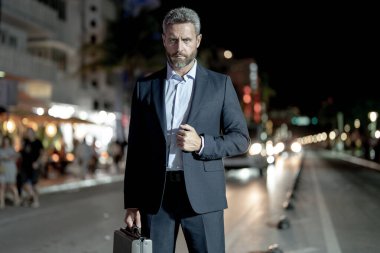 Gece vakti evrak çantalı bir iş adamı yürüyor. Şehirde takım elbiseli zengin bir adam. Lüks arabanın yanında bir iş adamı. Şehir ışıklarının yanında yürüyen bir iş adamı. İş stili. Lüks ortamda zengin bir işadamı
