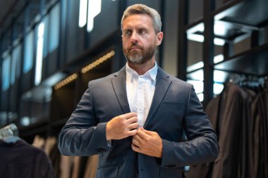 Takım elbiseli, butikte şık bir adam. Bir iş adamı mağazada şık bir takım elbise seçer. Modayı takip eden bir müşteri resmi kıyafet alırken ceketi inceler. Klasik kıyafetli çekici bir manken.