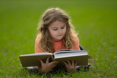 Çimlerin üzerinde kitap okuyan bir çocuk. Çocuk açık havada kitap okumaktan zevk alıyor. Güneşli bir günde çayırda kitapla ders çalışan bir çocuk. Doğada öğrenme. Öğrenci çocuk kitap okuyor. Çocuk açık havada öğreniyor