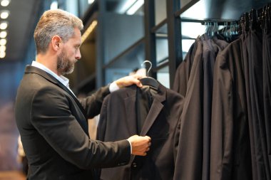 Olgun bir adam kıyafeti için mükemmel bir takım seçer. İyi giyimli bir erkek model bir butikte poz veriyor. Perakende satış asistanı şık bir adamın mükemmel takımı bulmasına yardım eder. Mağaza müdürü şık bir takım elbise ayarlar.