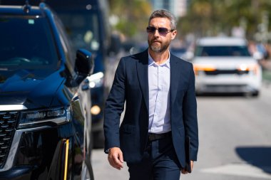 Lüks bir arabanın yanında zengin bir iş adamı. Takım elbiseli zengin bir iş adamı. Şehirdeki başarılı zengin iş adamı. Kendine güvenen bir iş adamı. Başarı işi. İş adamı sokakta yürüyor.