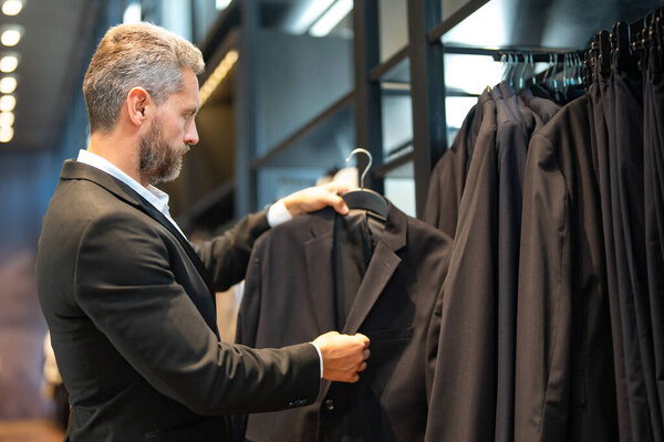 Зрелый мужчина выбирает идеальный костюм для своего наряда. Хорошо одетый мужчина позирует в бутике костюмов. Помощник продавца помогает стильному человеку найти идеальный костюм. Менеджер магазина организует элегантный костюм