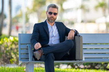 Kendine güvenen bir iş adamı bankta dizüstü bilgisayarı ve parktaki telefonuyla oturuyor. Zengin iş adamı. Olgun iş adamı tatilin tadını çıkarıyor. İş adamı parktaki bankta mola veriyor. Yaz işleri.