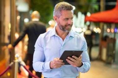 Gece sokakta tablet kullanan bir iş adamı. İş adamı dışarıda dijital tablet üzerinde çalışmayı planlıyor. Neon şehir ışıkları altında bir tablet üzerinde gezinen bir iş adamı. Serbest çalışan bir iş adamı. Gece işi.