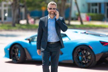 Lüks bir arabanın yanında zengin bir işadamı. Açık havada başarılı bir iş adamı. Zarif zengin iş adamı. Pahalı arabası olan bir patron. Premium bir arabanın önünde duran bir iş adamı. İş başarısı
