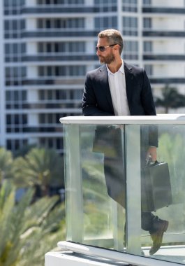 Şehir manzaralı bir balkonda duran bir iş adamı. Başarılı bir iş adamı yazın açık havada eğleniyor. Takım elbiseli bir iş adamı. İş başarısı. Şehir manzarası geçmişi olan bir iş adamı