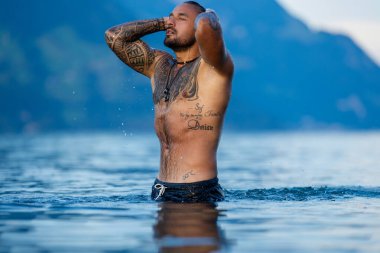 Seksi yaz erkeği. Kaslı vücut. Sıcak yaz. Gölde yüzen adam. Çıplak erkek vücudu. Adam yaz doğasında suya su sıçratıyor. Yaz plajında poz veren formda bir erkek model. Dağdaki gölde su sıçratan güçlü adam.