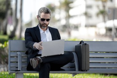 Kendine güvenen bir iş adamı bankta dizüstü bilgisayarı ve parktaki telefonuyla oturuyor. Başarılı bir iş adamı. Olgun iş adamı tatilin tadını çıkarıyor. İş adamı parktaki bankta mola veriyor. Yaz işleri.