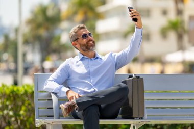 Kendine güvenen bir iş adamı bankta dizüstü bilgisayarı ve parktaki telefonuyla oturuyor. Başarılı bir iş adamı. Olgun iş adamı tatilin tadını çıkarıyor. İş adamı parktaki bankta mola veriyor. Yaz işleri.