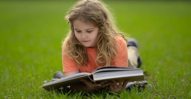 Çimlerin üzerinde kitap okuyan bir çocuk. Çocuk açık havada kitap okumaktan zevk alıyor. Güneşli bir günde çayırda kitapla ders çalışan bir çocuk. Doğada öğrenme. Öğrenci çocuk parkta kitap okuyor. Açık hava öğrenimi