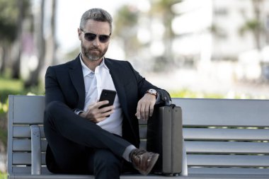 Kendine güvenen işadamı bankta dizüstü bilgisayarı ve parktaki telefonuyla oturuyor. Zengin iş adamı. Olgun iş adamı tatilin tadını çıkarıyor. İş adamı parktaki bankta mola veriyor. Kentsel iş