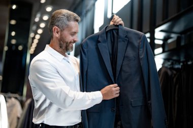 Takım elbiseli, butikte şık bir adam. Bir iş adamı mağazada şık bir takım elbise seçer. Modayı takip eden bir müşteri resmi kıyafet alırken ceketi inceler. Klasik kıyafetli çekici bir manken.