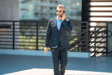 İş adamı telefonda konuşuyor ve yürüyor. Kendine güvenen bir iş adamı dışarıda telefonla konuşuyor. İş telefonu konuşması. Şirket lideri telefonda konuşmakla meşgul. Takım elbiseli zengin iş adamı. İş çağrısı