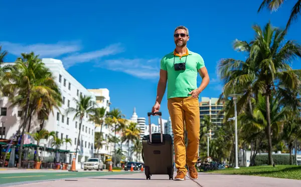 Bir gezgin Miami 'nin güneşli sokaklarında yürüyor. Bir erkek gezgin, tropik bir şehirde yaz tatilinin tadını çıkarır. Bavullu bir gezgin turist şehir manzarası. İnsan gezgin şehirde yürür