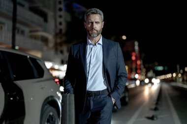 Gece vakti evrak çantalı bir iş adamı yürüyor. Şehirde takım elbiseli zengin bir adam. Lüks arabanın yanında bir iş adamı. Şehir ışıklarının yanında yürüyen bir iş adamı. İş stili. Lüks ortamda zengin bir işadamı