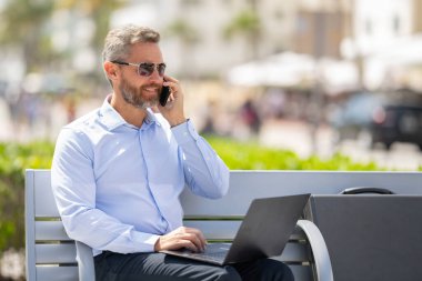 Kendine güvenen işadamı bankta dizüstü bilgisayarı ve parktaki telefonuyla oturuyor. Zengin iş adamı. Olgun iş adamı tatilin tadını çıkarıyor. İş adamı parktaki bankta mola veriyor. Yaz işleri.