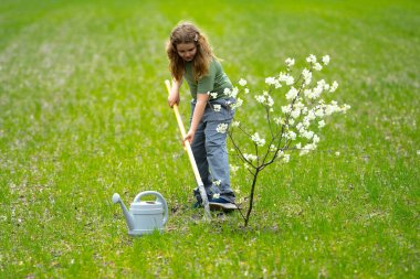 Çocuk dışarıda ağaç dikiyor. Bitkilerin bahar etkinliği. Çocuk kürek ve su bidonuyla çalışıyor. Çocuk meyve ağacı dikmiş. Bahçıvan çocuklar. Çocuk ağaç dikmek ve onunla ilgilenmek. Bahar otlağında küçük çiftçi