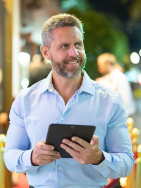 Gece sokakta tablet kullanan bir iş adamı. İş adamı dışarıda tablet üzerinde çalışmayı planlıyor. İşadamı neon şehir ışıkları altında bir tablet üzerinde geziniyor. Serbest çalışan bir iş adamı. Gece işi.