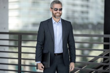 Başarılı bir iş adamı dışarıda dizüstü bilgisayarla dolaşıyor. Kendine güvenen iş adamı dizüstü bilgisayarı dışarıda tutuyor. Takım elbiseli, olgun, serbest çalışan bir iş adamı. Takım elbiseli zengin iş adamı