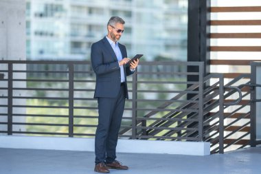 Açık havada tabletle başarılı bir iş adamı. Kendine güvenen iş adamı dışarıda tablet kullanıyor. Olgun iş adamı finans verilerini kontrol ediyor. Zengin iş adamı bankacı tabletteki sözleşmeyi gözden geçiriyor