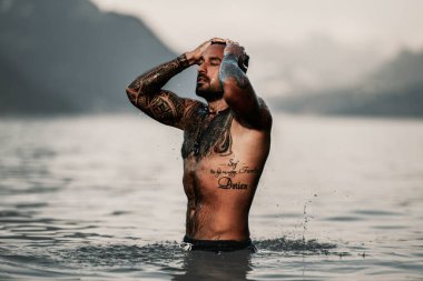 Seksi yaz erkeği. Suda ıslak kaslı erkek vücudu. Çıplak seksi erkek vücudu. İnsan yaz doğasında suya su sıçratır. Yaz plajında poz veren formda bir adam. Dağdaki gölde su sıçratan güçlü adam.