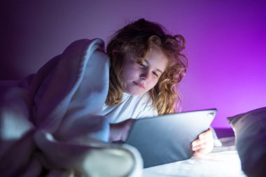 Çocuk yatakta uzanıyor ve karanlık odada tablet üzerinde internette sörf yapıyor. Geceleri Tablet okuyan bir çocuk. Yatmadan önce pijamalı bir çocuk tablet kullanıyor. Çocuk tablet aletleri kullanıyor.