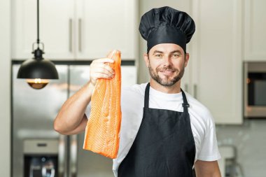 Mutfakta taze somon balığı hazırlayan gülümseyen adamın portresi. Yakışıklı, neşeli şef çiğ balık somonu hazırlıyor. Sağlıklı yiyecekler, yemek pişirme konsepti