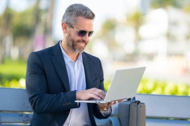 Kendine güvenen bir iş adamı bankta dizüstü bilgisayarı ve parktaki telefonuyla oturuyor. Başarılı bir iş adamı. Olgun iş adamı tatilin tadını çıkarıyor. İş adamı parktaki bankta mola veriyor. Yaz işleri.