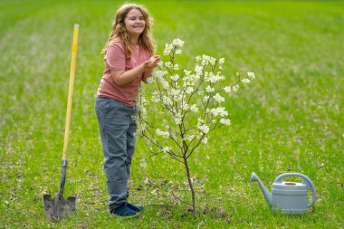 Bahçeye çocuk dikme ve sulama ağacı. Ağaç dikmenin bahar etkinliği. Kürek ve su bidonlarıyla çocuk işi. Çocuk meyve ağacı dikiyor. Bahçıvan çocuklar. Çocuk yetiştirme ve ağaç bakımı