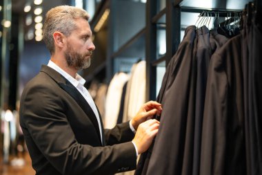 Bir adam butikte alışveriş yaparken klasik bir terzi kıyafeti giyer. Başarılı bir iş adamı için klasik bir kıyafet. Takım elbiseli modaya uygun bir mağaza. Takım elbise seçen bir iş adamı. Bir erkek resmi etkinlik için takım elbise seçer.