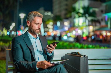 Takım elbiseli bir adam gece telefonda konuşuyor. İş adamı sokakta bir telefon görüşmesi yapıyor. Tabletli ve telefonlu bir iş adamı. İş adamı telefonda bankta bekliyor. Gece işi.