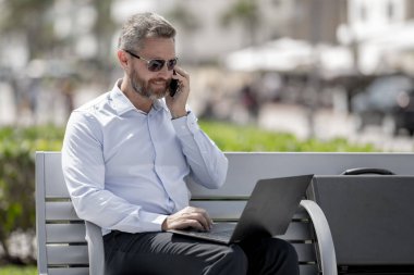 Kendine güvenen işadamı bankta dizüstü bilgisayarı ve parktaki telefonuyla oturuyor. Başarılı bir iş adamı. Olgun iş adamı tatilin tadını çıkarıyor. İş adamı parktaki bankta mola veriyor. Yaz işleri.