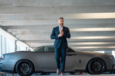 Lüks bir arabanın yanında zengin bir iş adamı. Takım elbiseli zengin bir iş adamı. Şehirdeki başarılı zengin iş adamı. Kendine güvenen bir iş adamı. Başarı işi ve zenginlik. İş adamı sokakta yürüyor