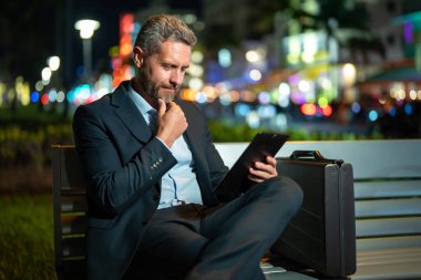 Takım elbiseli bir adam dışarıda tablet üzerinde çalışıyor. Tabletli bir iş adamı. Gece sokağında tabletli bir iş adamı. Tablet kullanan bir işadamı, bankta oturur. Gece işi.