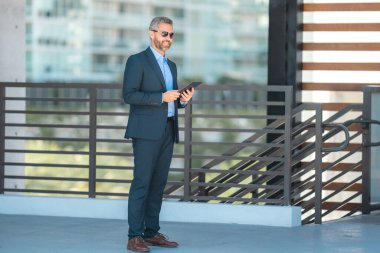 Açık havada tabletle başarılı bir iş adamı. Kendine güvenen bir iş adamı dışarıda tablet kullanıyor. Olgun iş adamı girişimci finans verilerini kontrol eder. Zengin bankacı tabletteki sözleşmeyi gözden geçiriyor