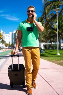 Miami sokaklarında kamera ve bavul taşıyan bir gezgin. Bir erkek gezgin, tropik bir şehirde yaz tatilinin tadını çıkarır. Yaz şehrinde bavullu bir turist. İnsan gezgin şehirde yürür