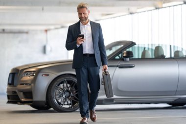 Dışarıdaki iş adamı. Zengin iş adamı başarının tadını çıkarıyor. Şık takım elbiseli lüks iş adamı. Pahalı arabanın önündeki zengin adam. Lüks iş seyahati. Klasik takım elbiseli, kendinden emin zengin bir iş adamı.