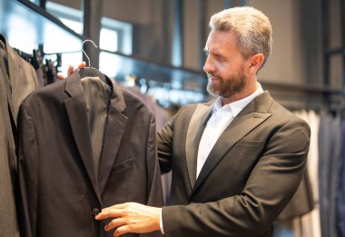 Olgun bir adam kıyafeti için mükemmel bir takım seçer. İyi giyimli bir erkek model bir butikte poz veriyor. Perakende satış asistanı şık bir adamın mükemmel takımı bulmasına yardım eder. Mağaza müdürü şık bir takım elbise ayarlar.